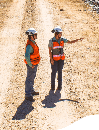 Duas empregadas Vale em um chão de areia. Elas estão de colete laranja e capacetes. Uma aponta para o horizonte e a outra olha na mesma direção.