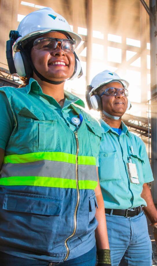 Dois empregados da Vale – um homem e uma mulher – parados de pé um do lado do outro em uma área de operações. Os dois estão sorrindo e usam roupas verdes, óculos de proteção, protetores de ouvido e capacete branco com logotipo da Vale