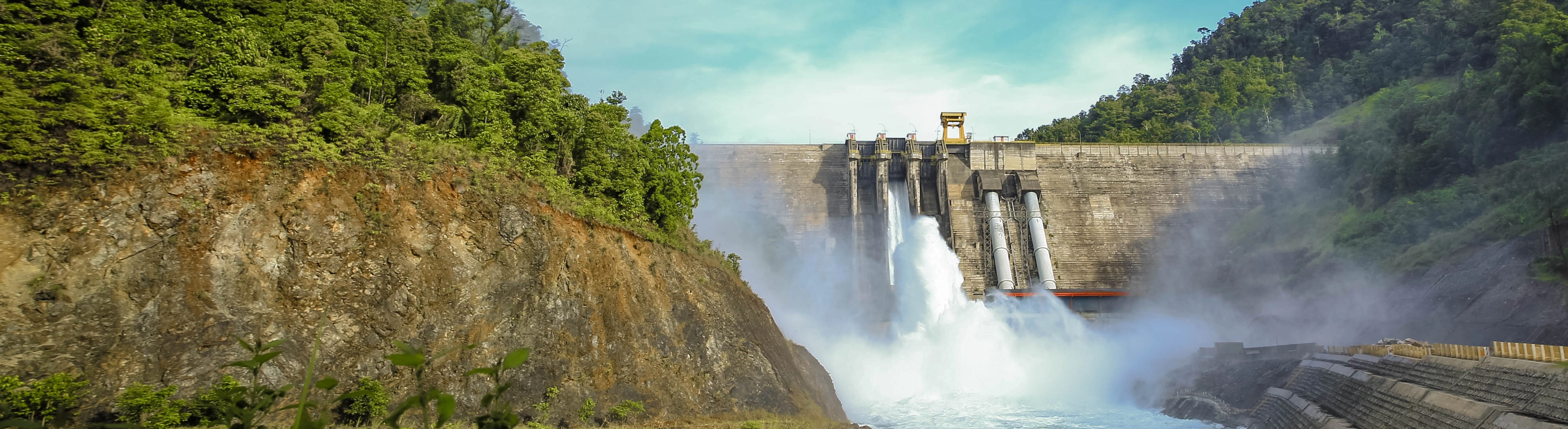 Hydroelectric power plant. There are large, tall concrete structures in most of the image, and water below. In the foreground is a mountain