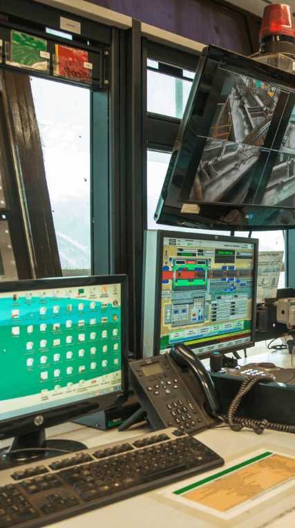 Two Vale employees, a bald man with a beard and a blond woman with straight hair, are in a control room, full of monitors. The man is standing, pointing at a screen and the woman is sitting, paying attention.