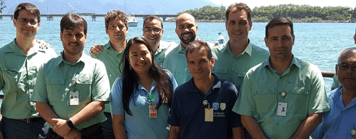 Foto de oito homens e uma mulher com mar e uma ponte ao fundo em um dia de sol. Sete homens estão de uniforme da Vale, camisa de botão verde de manga curta, calça jeans e crachá. Um dos homens está de camisa polo azul marinho, calça jeans e crachá. A mulher está de blusa polo azul clara, calça jeans e crachá