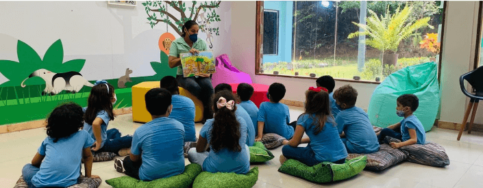 Some children look at Vale employee, while she is showing a book. The children are sitting on cushions and wear blue uniforms.