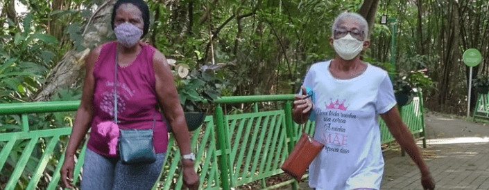 Duas senhoras caminham por um local arborizados. Ambas usam máscara de proteção.