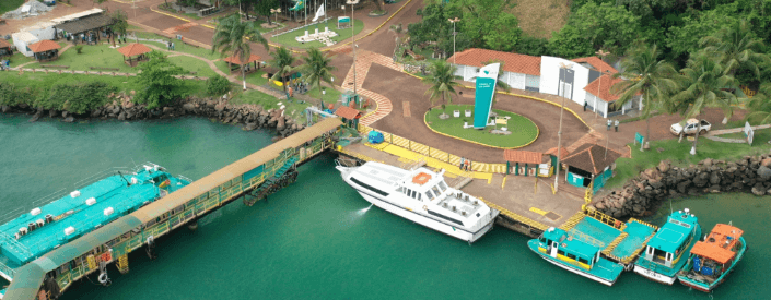 Aerial image of the Guaíba Island terminal. At the top of the image, we see an area of land on the left. On the right, a mountainous area with vegetation. In the middle there are several concrete structures, and below, you can see the water, a moored motorboat, and a kind of bridge.