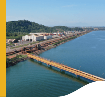 Aerial image of the Itaguaí port. You can see a bridge under the water and, in the background, some sheds and a mountainous area.