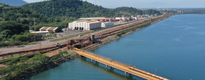 Aerial image of the Itaguaí port. You can see a bridge under the water and, in the background, some sheds and a mountainous area.