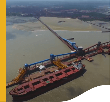 Aerial photo of the Ponta Madeira terminal. In addition to a large docked cargo ship, you can see large, belt-like equipment. The place is surrounded by brown colored water, and in the background you can see vegetation and some buildings.