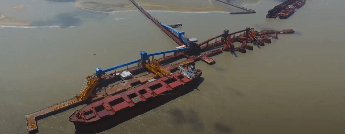 Aerial photo of the Ponta Madeira terminal. In addition to a large docked cargo ship, you can see large, belt-like equipment. The place is surrounded by brown colored water, and in the background you can see vegetation and some buildings.