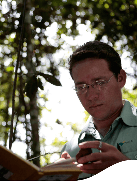 Foto de um homem com árvores ao fundo, olhando para o caderno. Ele está usando camisa de botões verde da Vale e óculos de grau.