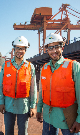 Photo of two men smiling in an operation area. They are both wearing green Vale button-up shirts, orange vests, white helmets with Vale logos, and goggles. One of them is holding a clipboard in his hand.