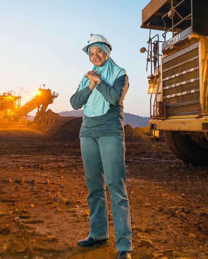 Photo of a woman in an operation area, with iron structures in the background. The woman is wearing a long-sleeved shirt, a veil covering her head and neck, a white helmet with a Vale logo.