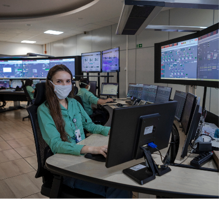 Photo of a woman sitting on a chair working on a computer in an office with several monitors and screens in the background. She has long dark hair and is wearing a uniform, a green button-up blouse, badge, and face mask.