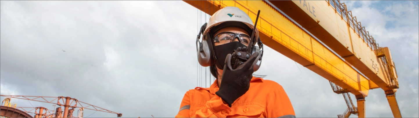 Photo of a woman in an operation area talking on a radio. She is wearing a Vale uniform, orange shirt, black glove, face mask, goggles, white helmet with Vale logo, and ear protection.