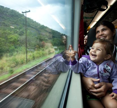 A woman and a child in her arms look out the train window and smile at the landscape, composed of vegetation.