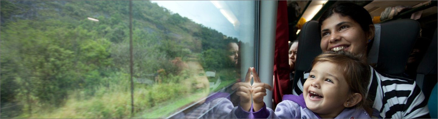 Uma mulher e uma criança  de colo olham pela janela do trem e sorrirem para paisagem, composta por vegetação.