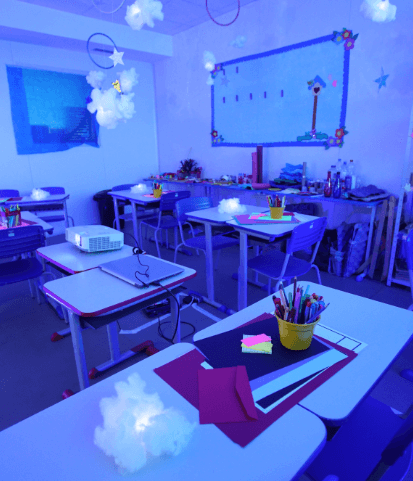 A classroom with the light off and some points of light scattered around the desks, where there are colorful papers and pencils as well.