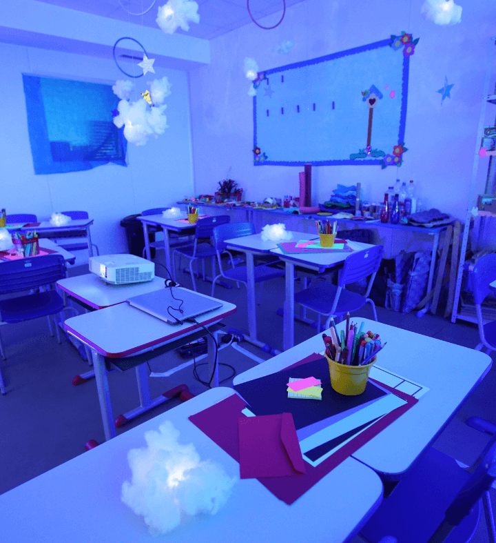 A classroom with the light off and some points of light scattered around the desks, where there are colorful papers and pencils as well.