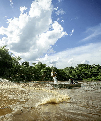 In a river surrounded by dense vegetation, two men wearing life jackets are inside a small boat.   One of the men is standing and casting a net in the water.