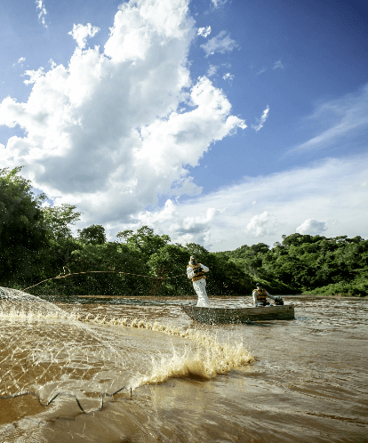 Em um rio, cercado por densa vegetação, dois homens de colete salva-vidas estão dentro de um pequeno barco. Um dos homens está de pé e lança uma rede sob a água.
