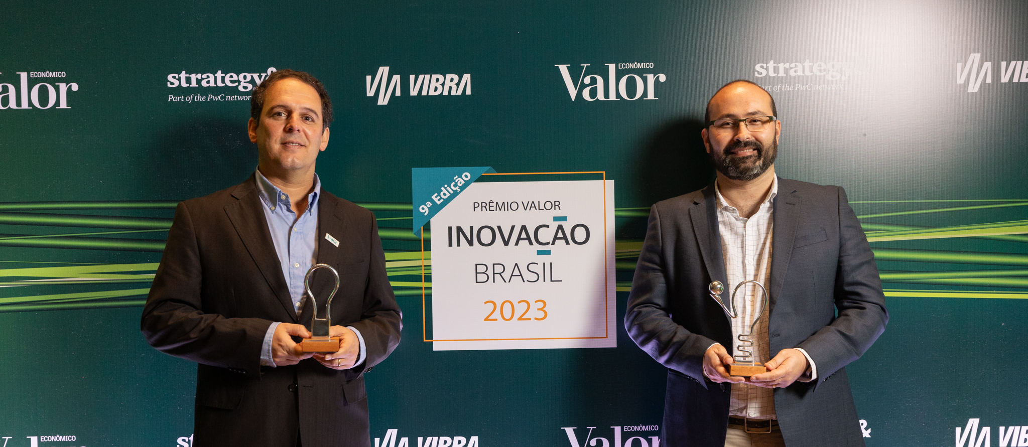 Dois homens usando roupas sociais seguram prêmios em frente a um painel onde está escrito: 9ª edição. Prêmio Valor Inovação Brasil 2023