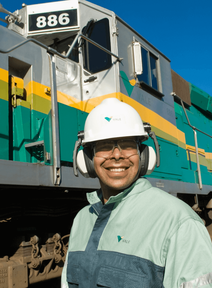 Um homem usa uma camisa em um tom de verde claro, capacete branco, óculos de proteção e proteção nos ouvidos. Ele está sorrindo em um lugar aberto e atrás deles tem um trem da Vale verde, amarelo e cinza.