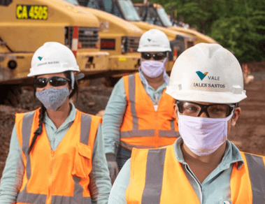 Foto da altura do peito até a cabeça de três empregados da Vale, dois homens e uma mulher. Os três estão com uniforme verde da Vale, colete laranja e cinza, óculos, capacete e máscara.