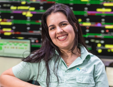 Foto de uma mulher em um escritório e vários monitores ao fundo. Ela está apoiada em um balcão e está usando camisa verde de botões com logo da Vale, cabelo comprido solt liso e está sorrindo.