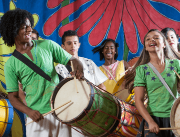 Jovens tocam tambores e sorriem, como se estivessem cantando. Atrás há um tecido estampado com as cores azul, vermelho, amarelo e verde.