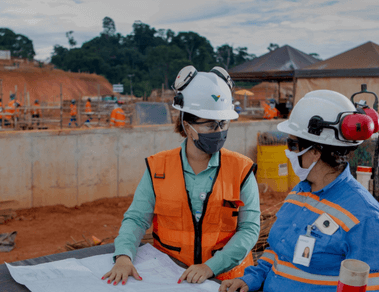 Foto de duas mulheres conversando e apontando para um papel grande e ao fundo uma área com obra com tendas, terra, cimento e pessoas trabalhando. Uma das mulheres está usando camisa verde de botões da Vale, colete laranja, óculos de proteção, máscara e capacete. Já a outra mulher está usando uma camisa azul com linhas fluorescentes, máscara, óculos de proteção e capacete.