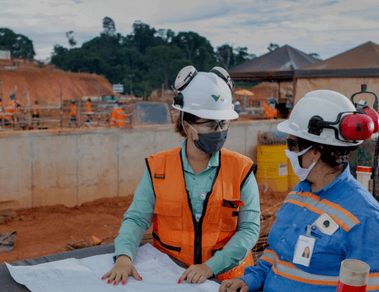 Photo of two women talking and pointing to a big paper, and in the background, an area with construction works with tents, earth, cement and people working. One of the women is wearing a green Vale button-down shirt, an orange vest, goggles, a mask and a helmet. The other woman is wearing a blue shirt with fluorescent lines, a mask, goggles and a helmet.