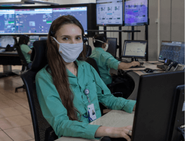 Photo of a woman sitting on a chair working on a computer in an office with several monitors and screens in the background. She has long dark hair and is wearing a uniform, a green button-up shirt, badge, and face mask.