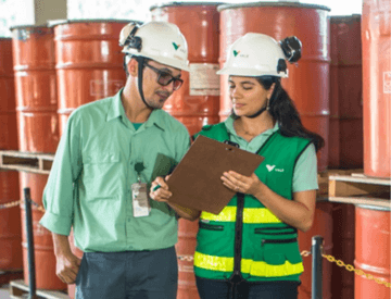 A man and a woman standing side by side looking at a clipboard. They wear jeans, green uniform shirts and helmets with Vale´s logo. The woman wears a dark green vest with lime green details. In the background, there are several stacked containers.