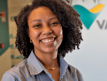 Mulher negra parada em um espaço de escritório, usando camisa cinza e sorrindo.