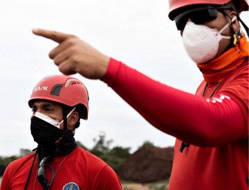 Two firefighters are looking at the horizon and one of them is pointing straight ahead. They are wearing protective masks and helmets.