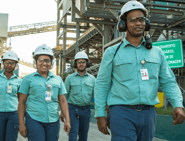 Empregados e empregadas andando lado a lado em um espaço de operações. Todos usam uniforme verde, óculos de proteção, protetores de ouvido e capacete branco com logotipo da Vale.