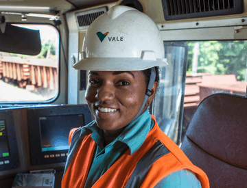 Mulher negra sorrindo dentro de um veículo. Ela usa uniforme verde da Vale, colete laranja e capacete branco com logotipo da empresa.