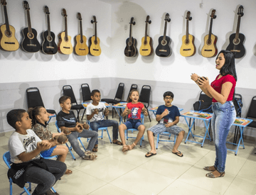 Sala de música com diversos violões pendurados na parede. Ao centro, há diversas crianças sentadas em círculos prestando atenção em uma mulher que gesticula.