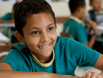 Um menino, dentro de uma sala de aula, pinta uma folha com giz de cera. Ele usa aparelho nos dentes e veste um uniforme verde e amarelo.