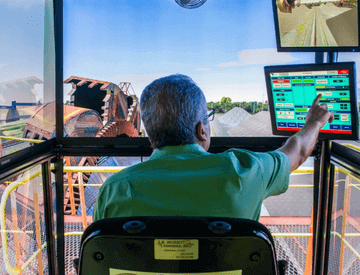 Empregado Vale, sentado de costas para foto, operando remotamente um equipamento. Ele está tocando em uma das duas telas que há em sua frente. Ao fundo, é possível ver um maquinário grande.