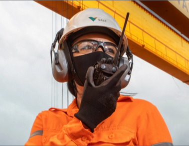Photo of a woman in an operation area talking on a radio. She is wearing a Vale uniform, orange shirt, black glove, face mask, goggles, white helmet with Vale logo, and ear protection.