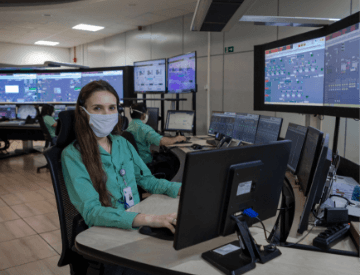 Foto de uma mulher sentada em uma cadeira mexendo no computador em um escritório com vários monitores e telas ao fundo. Ela tem cabelo longo e escuro e está usando uniforme, blusa de botões verde, crachá e máscara no rosto.