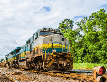 Trem de carga Vale trafegando por um trilho cheio de pedras. Oo fundo é possível ver algumas árvores.