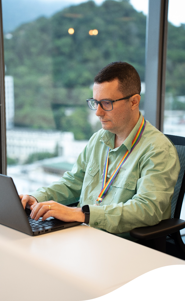 Um homem está sentado, digitando em um notebook. Ele usa óculos, uniforme Vale e um cordão com as cores LGBT no pescoço.