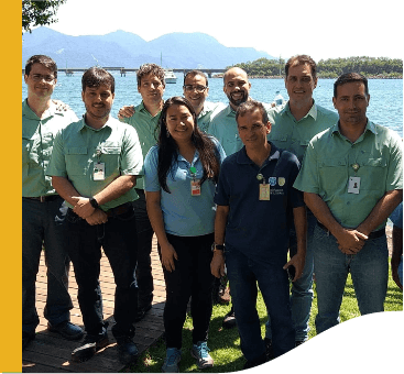 Foto de oito homens e uma mulher com mar e uma ponte ao fundo em um dia de sol. Sete homens estão de uniforme da Vale, camisa de botão verde de manga curta, calça jeans e crachá. Um dos homens está de camisa polo azul marinho, calça jeans e crachá. A mulher está de blusa polo azul clara, calça jeans e crachá